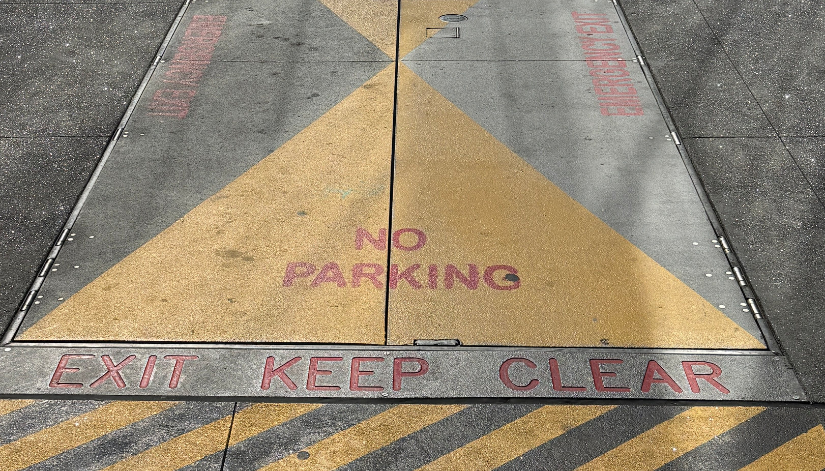 A painted yellow “EXIT—KEEP CLEAR / NO PARKING” hatch on a downtown sidewalk marks a sidewalk-level emergency egress panel; a pay-to-park kiosk and curbside stalls sit immediately adjacent, creating risk that a parked vehicle could block the exit.
